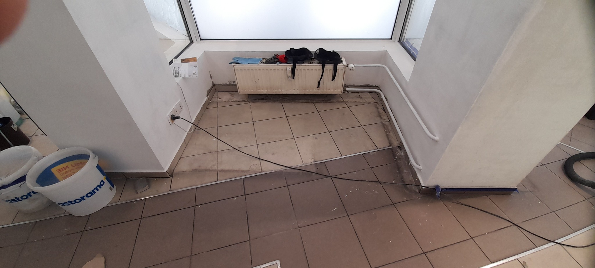 Wnętrze w trakcie prac wykończeniowych: widoczne dwa białe wiadra z farbą Castorama, zabezpieczone listwy przypodłogowe niebieską taśmą malarską, grzejnik z suszącymi się rękawicami roboczymi...