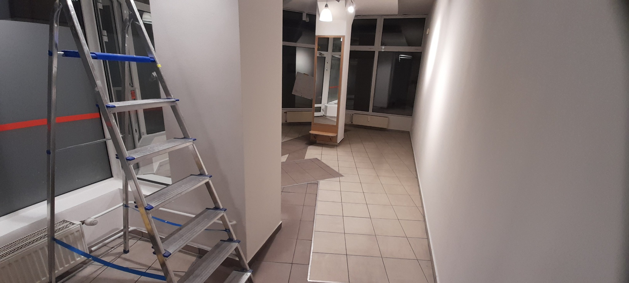 Wnętrze w trakcie malowania: aluminiowa drabina z niebieskimi zabezpieczeniami stoi przy świeżo pomalowanej, jasnoszarej ścianie, widoczny fragment grzejnika i okna z czerwoną linią, w tle korytarz...