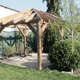 Drewniana konstrukcja z czterema słupami i zadaszeniem w trakcie budowy na przygotowanym podłożu z płyt, narzędzia leżą obok, w tle zielony płot i roślinność.