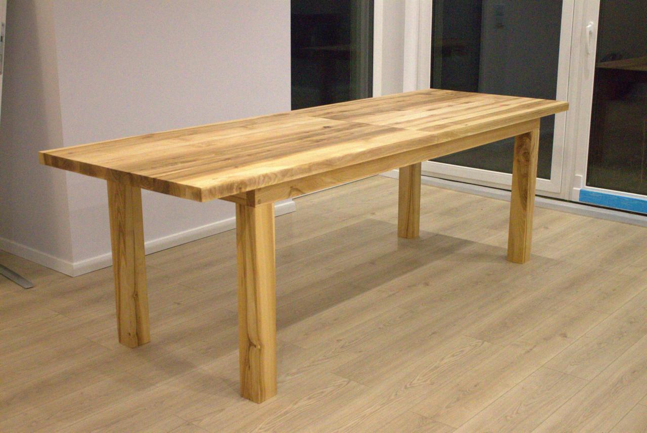 Prostokątny stół z jasnego drewna z widocznymi słojami, ustawiony na drewnianej podłodze, z białymi drzwiami balkonowymi w tle.