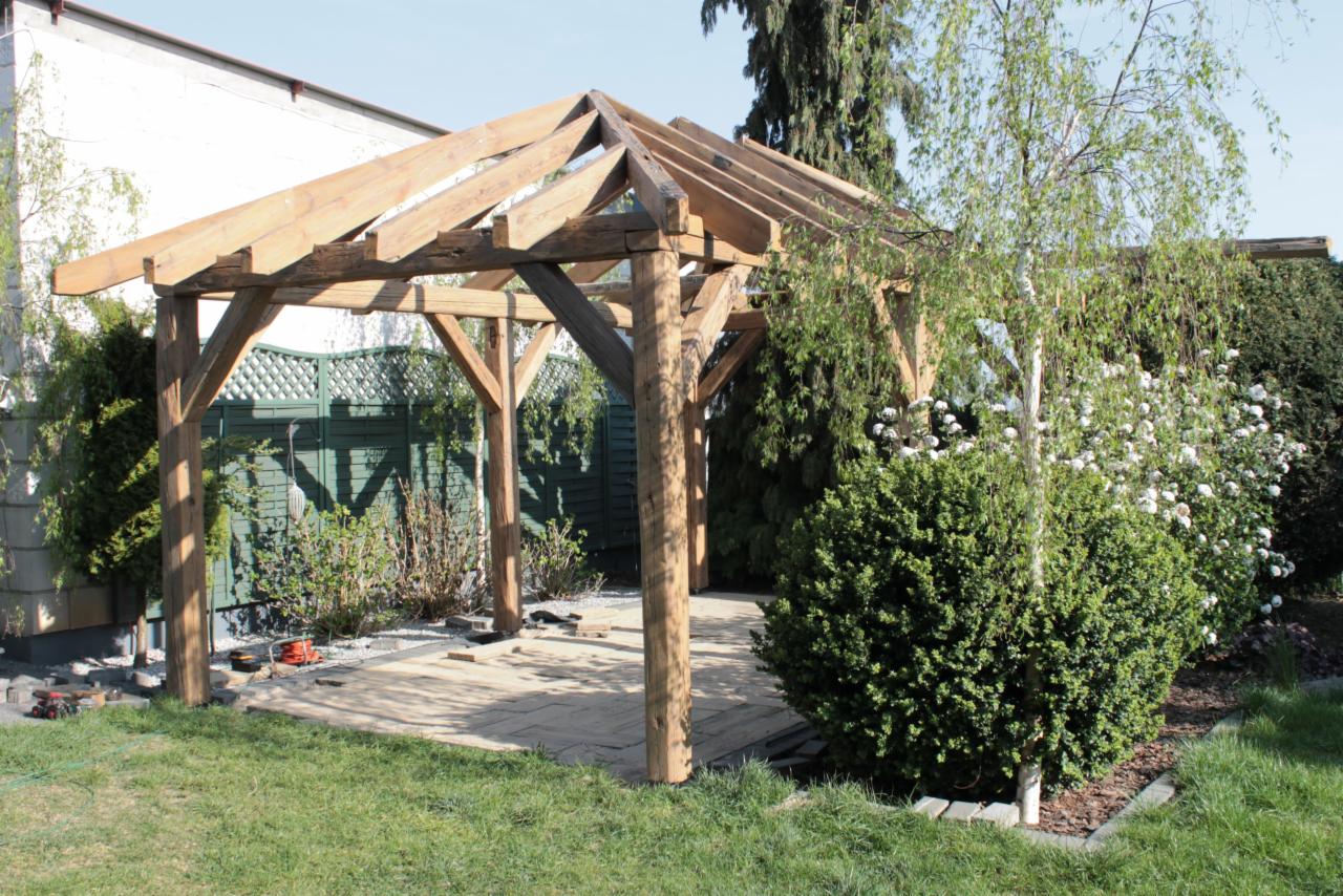 Drewniana konstrukcja z czterema słupami i zadaszeniem w trakcie budowy na przygotowanym podłożu z płyt, narzędzia leżą obok, w tle zielony płot i roślinność.