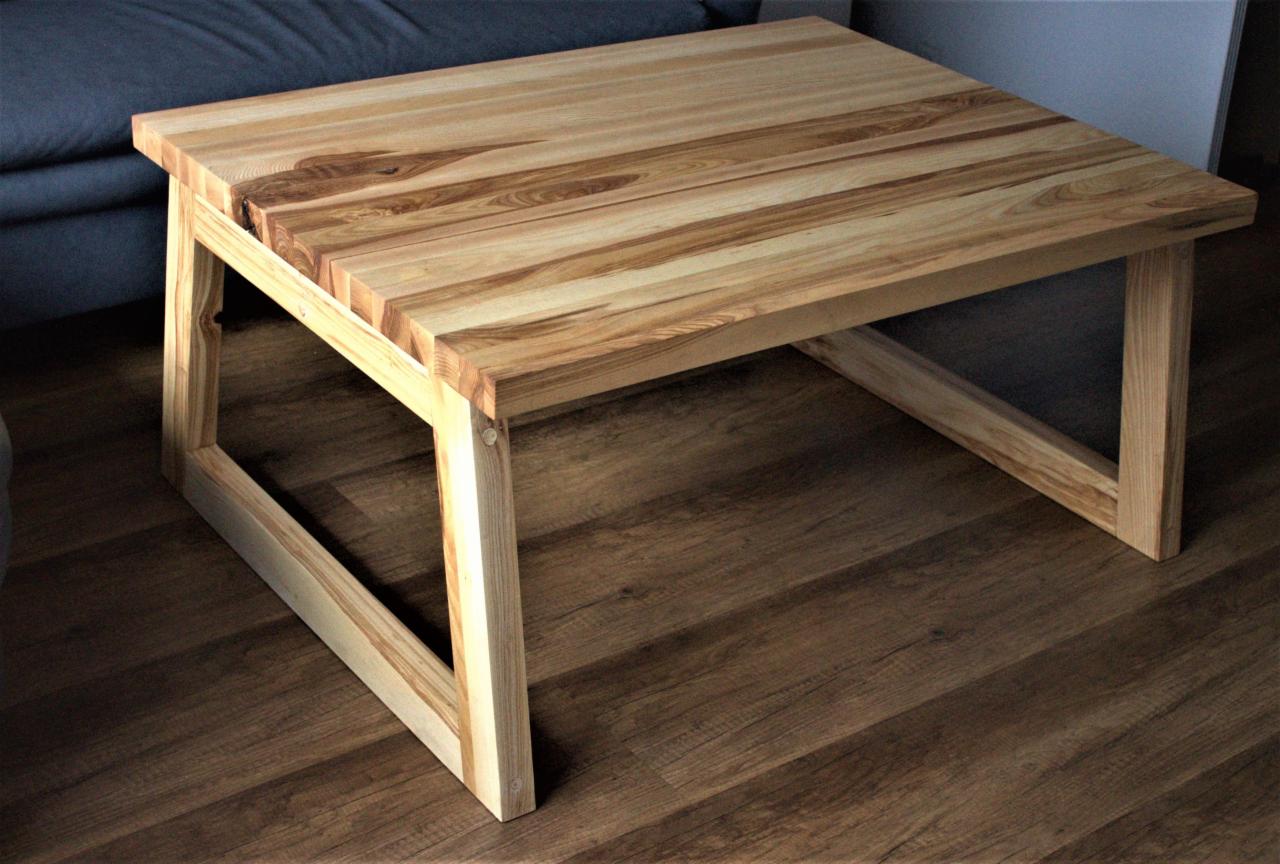 Niski stolik kawowy z jasnego drewna o wyraźnym usłojeniu, z nogami w kształcie litery 'A', na drewnianej podłodze, widoczny fragment szarej sofy w tle.