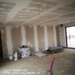 Pokój w trakcie prac wykończeniowych: ściany częściowo pokryte gładzią, widoczne płyty gipsowo-kartonowe, przewody elektryczne wystające z sufitu, paleta z materiałami budowlanymi i narzędzia...