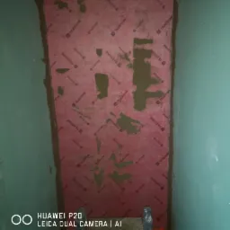 Przygotowanie ściany w łazience pod płytki: czerwona mata izolacyjna z nadrukiem 'TILESTONE' przyklejona do ściany z nałożonymi punktowo fragmentami zaprawy klejowej.