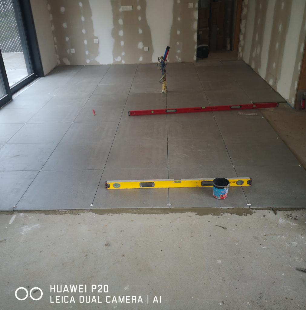 Ułożone szare płytki podłogowe w trakcie prac wykończeniowych, widoczne poziomice, wiadro z zaprawą i krzyżyki dystansowe, ściany przygotowane do malowania.