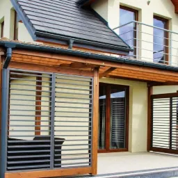 Zadaszony taras z drewnianą konstrukcją i metalowymi panelami żaluzjowymi, przylegający do domu z beżową elewacją i brązowym dachem.