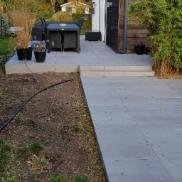 Szara, betonowa powierzchnia przydomowa z zestawem mebli ogrodowych i roślinami w donicach, widok na budynek z drewnianą elewacją i trawnik z fragmentami narzędzi ogrodniczych.