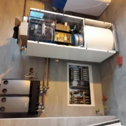 Wnętrze kotłowni z zamontowanym, otwartym kotłem gazowym z widocznymi elementami wewnętrznymi, zbiornikiem i modułem sterującym, obok dodatkowy moduł grzewczy i instalacja wnękowa, na szarej...