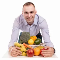 Uśmiechnięty mężczyzna w błękitnej koszuli prezentuje koszyk pełen świeżych owoców i warzyw na białym tle, w tym banany, jabłka, pomarańcze, awokado, brokuły i paprykę.