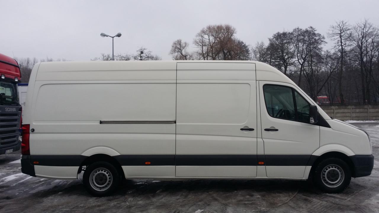 Biały, długi bus dostawczy marki Volkswagen Crafter z przesuwnymi drzwiami, zaparkowany na parkingu w pochmurny dzień, widoczny z boku, obok czerwonego samochodu ciężarowego marki Scania.