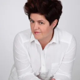 Portret kobiety w białej koszuli, opierającej się o kolumnę, z krótkimi brązowymi włosami i delikatnym uśmiechem.