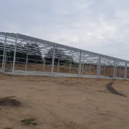 Szkielet konstrukcji stalowej hali w trakcie budowy, widoczne słupy i kratownice, teren piaszczysty, w tle drzewa.