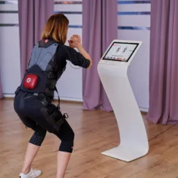 Kobieta w kombinezonie EMS ćwiczy przysiady przed ekranem dotykowym w jasnym pomieszczeniu z drewnianą podłogą.