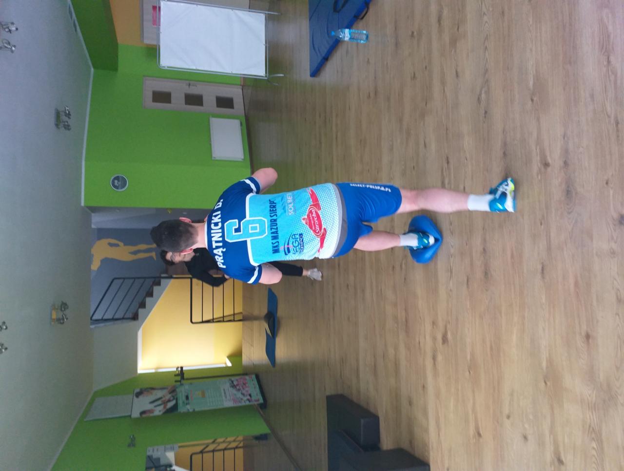 Mężczyzna w sportowym stroju wykonuje ćwiczenie na podłodze z użyciem niebieskich poduszek sensorycznych, w tle widoczna osoba i wyposażenie sali do ćwiczeń.