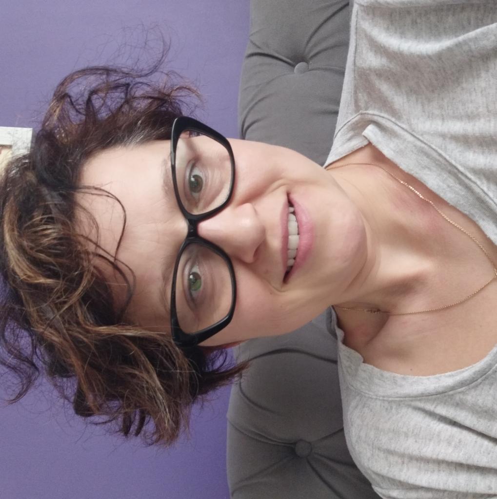 Portret uśmiechniętej kobiety w okularach, z brązowymi włosami i złotym łańcuszkiem, na tle fioletowej ściany i szarej tapicerki.