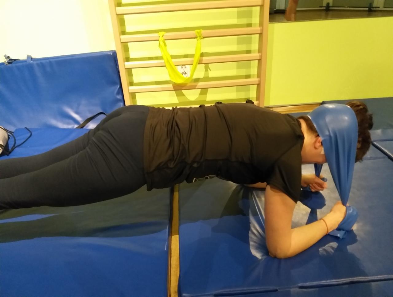 Osoba w czarnym stroju ćwiczy plank z niebieską gumą oporową na podłodze wyłożonej niebieską matą, drabinki gimnastyczne w tle.