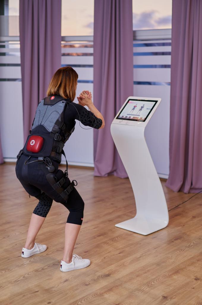 Kobieta w kombinezonie EMS ćwiczy przysiady przed ekranem dotykowym w jasnym pomieszczeniu z drewnianą podłogą.