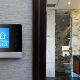 Panel sterowania inteligentnego domu zamontowany na ścianie, wyświetlający informacje o pogodzie, temperaturze, dacie i ikonach funkcji bezpieczeństwa, z widokiem na luksusową łazienkę...