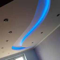 Nowoczesny sufit podwieszany z niebieskim oświetleniem LED w kształcie fali, punktowe lampy halogenowe i okno w tle.