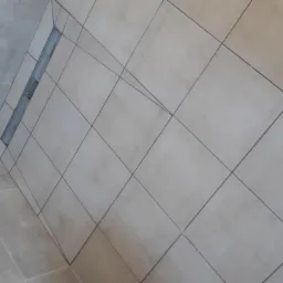 Beżowe płytki podłogowe ułożone ukośnie z odpływem liniowym w rogu łazienki.