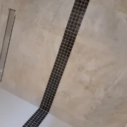 Ukośne ujęcie wykończonej podłogi prysznica z odpływem liniowym i pasem czarnej mozaiki na tle płytek imitujących marmur.