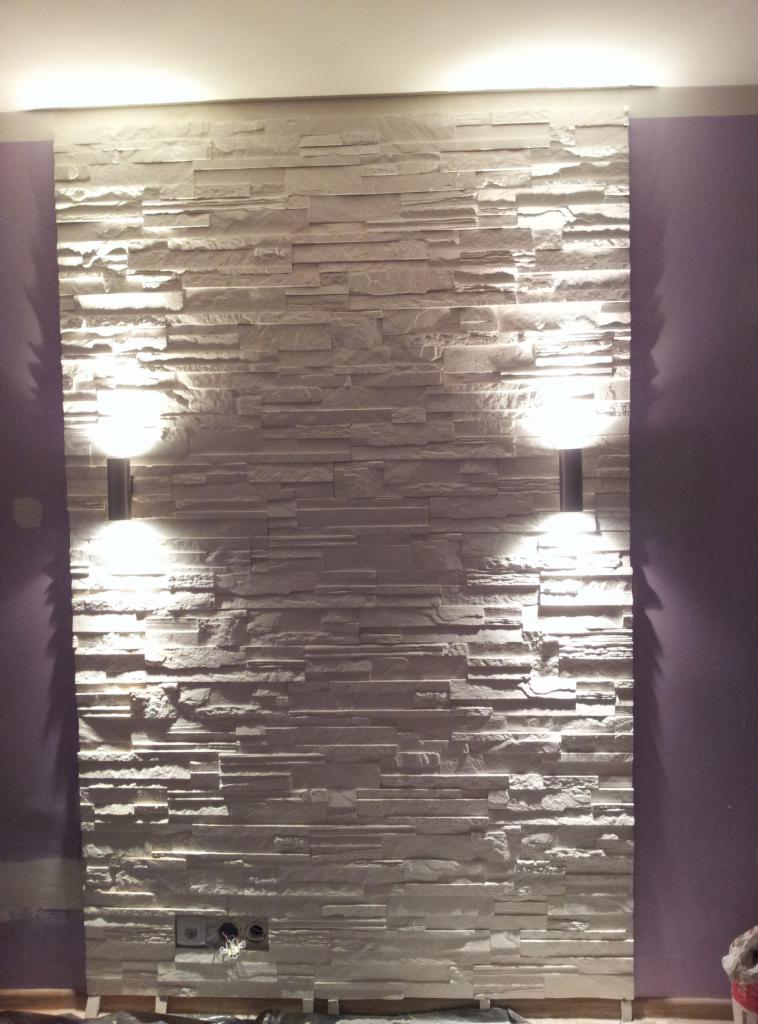 Ściana z dekoracyjnych, białych płytek imitujących kamień z oświetleniem punktowym na fioletowym tle ściany, widoczne gniazdka elektryczne.