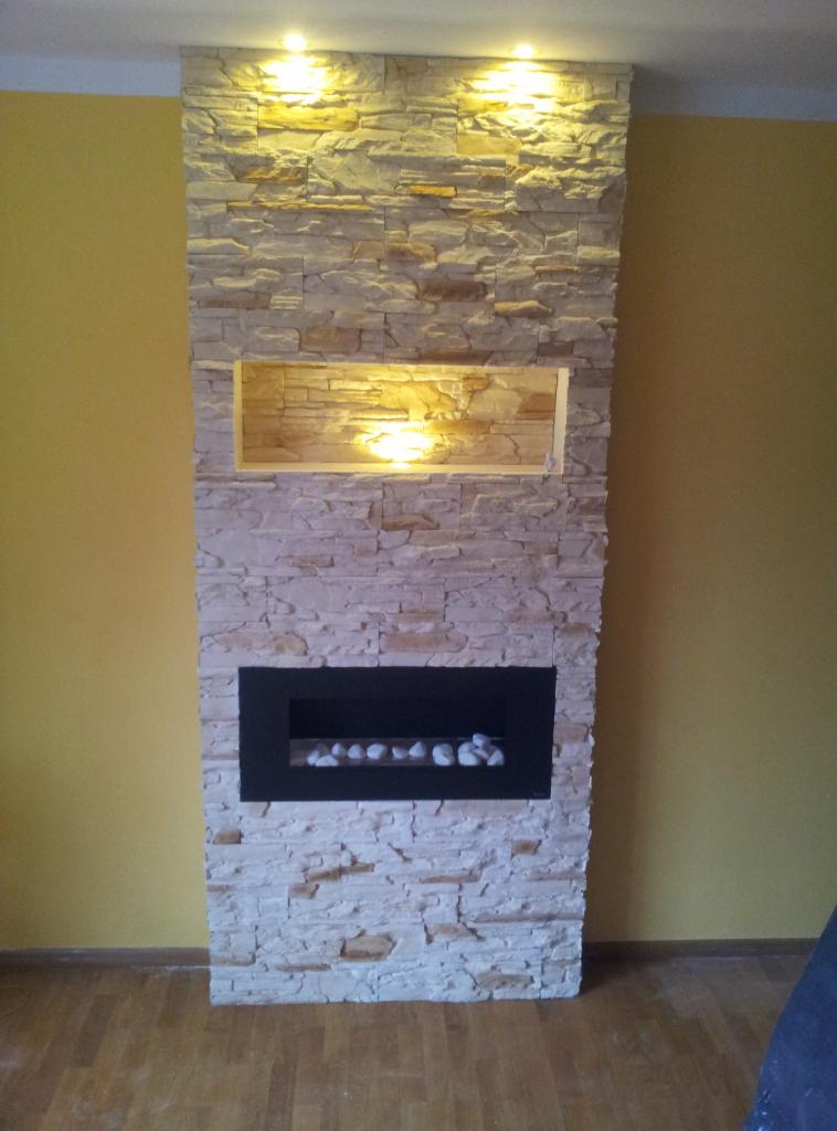 Ściana dekoracyjna z kamienia elewacyjnego z wnęką oświetleniową i czarnym biokominkiem z białymi kamieniami, na tle żółtej ściany i drewnianej podłogi.