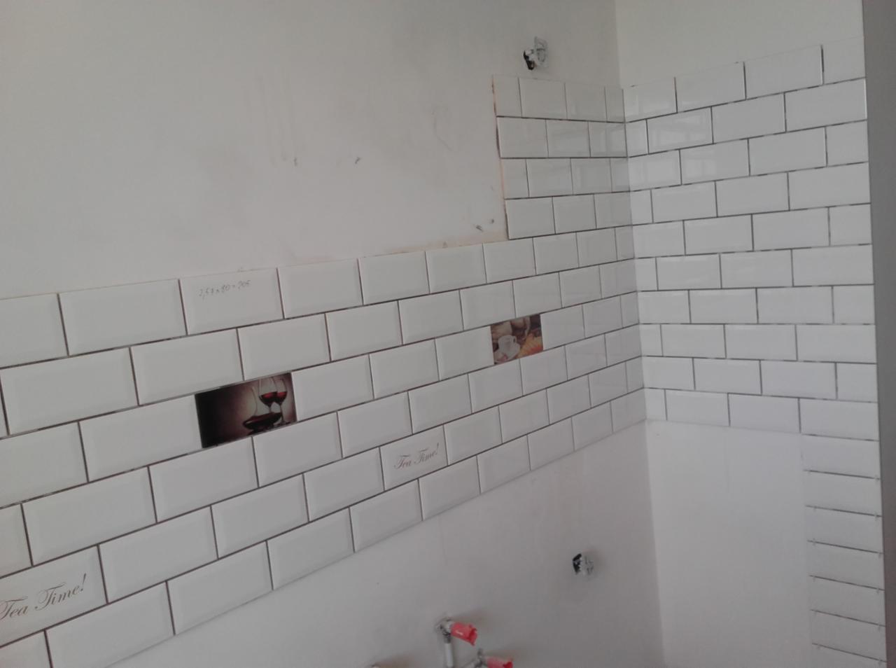 Białe, prostokątne płytki ułożone na ścianie w kuchni, niektóre z nadrukiem tematycznym, widoczne niedokończone prace przy montażu płytek.