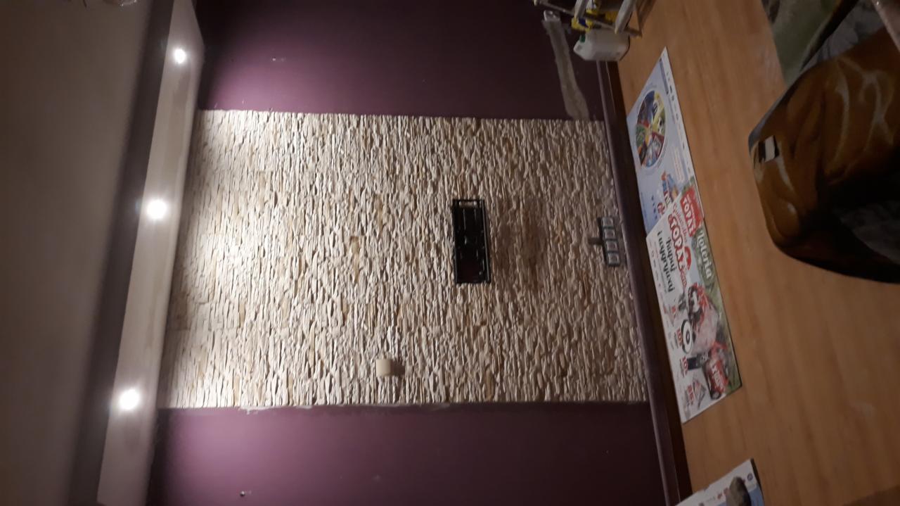 Ściana wyłożona dekoracyjnym kamieniem z zamontowanym uchwytem pod telewizor, dolna i górna część ściany pomalowana na fioletowo, oświetlenie punktowe w suficie.