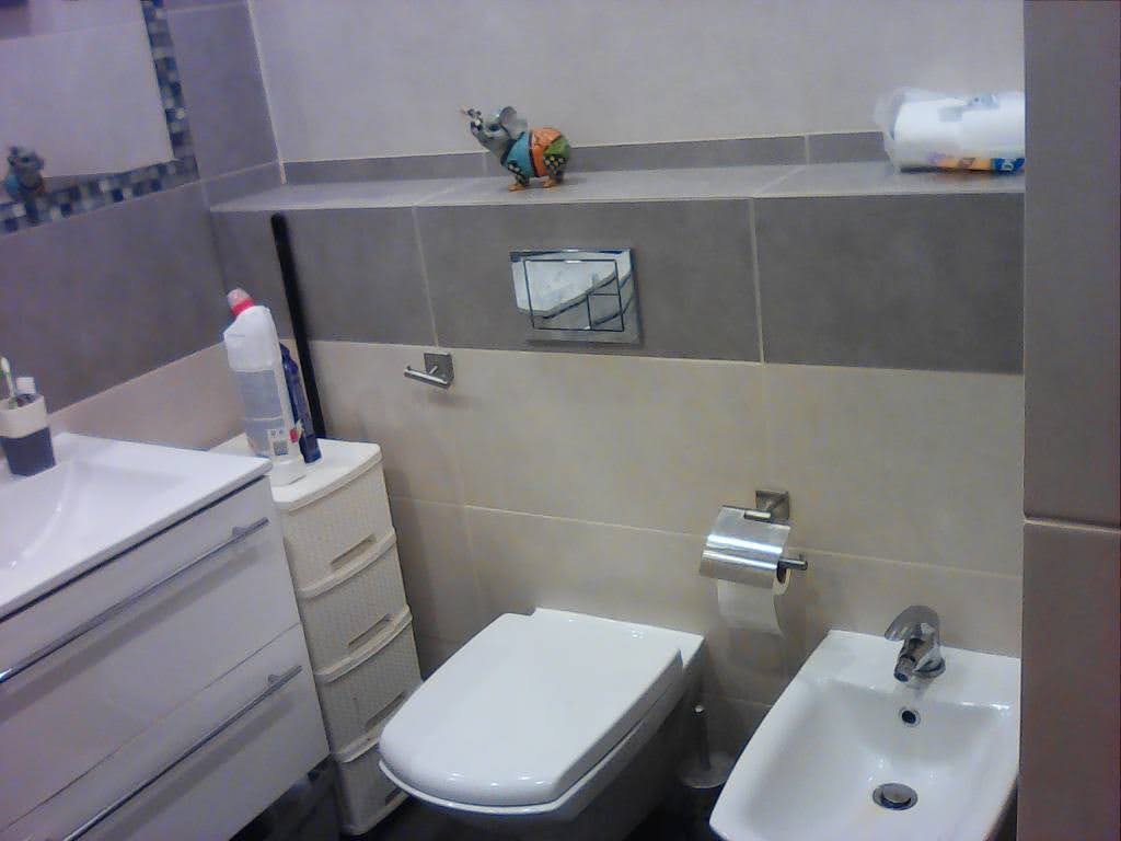 Wykończona łazienka z bidetem, toaletą i umywalką, wykończona płytkami w odcieniach beżu i szarości, z dekoracyjną figurką zwierzęcia na półce.