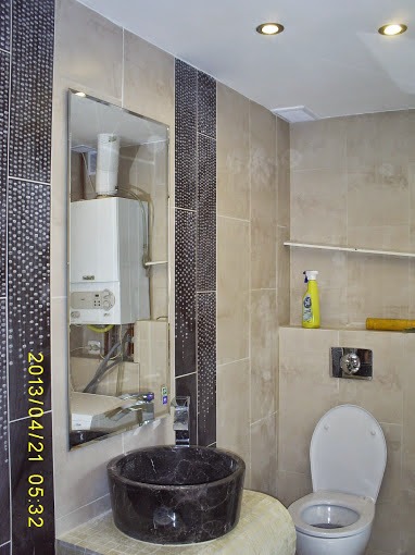 Wykończona łazienka z beżowymi płytkami, dekoracyjnymi pasami z mozaiki, okrągłą umywalką z ciemnego kamienia i widocznym w lustrze piecem gazowym.