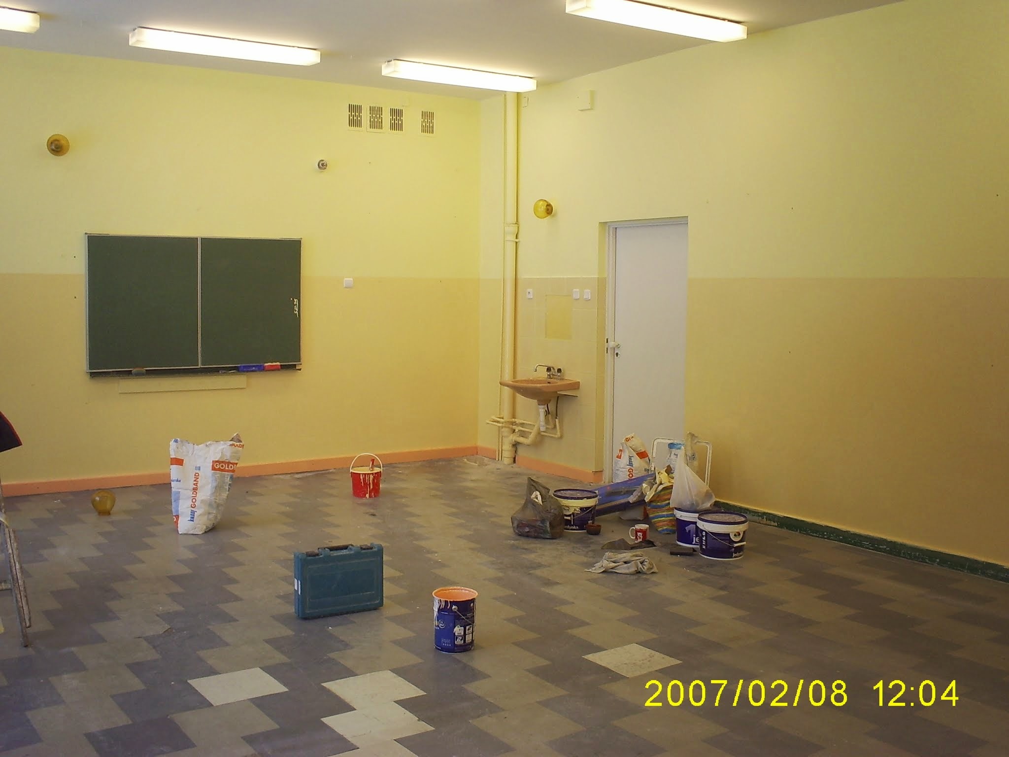 Pokój w trakcie malowania ścian na żółto, widoczne wiaderka z farbą, worek gładzi, kuweta malarska i niebieska skrzynka narzędziowa na podłodze z wzorem w zygzaki.