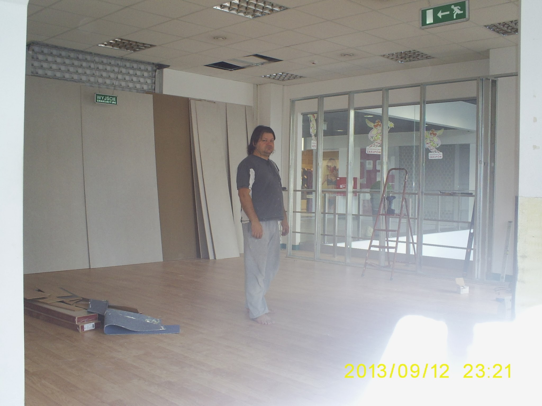 Mężczyzna stoi na nowo ułożonych panelach podłogowych w trakcie prac wykończeniowych w lokalu handlowym, widoczne płyty gipsowe oparte o ścianę i drabina.