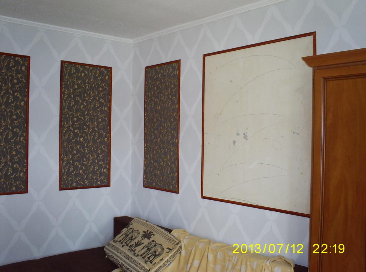 Pokój z dekoracyjną tapetą w romby, trzema panelami ściennymi z wzorzystą tapetą w drewnianych ramach oraz przygotowaną ścianą do malowania, widoczny narożnik sofy z poduszką z motywem słoni.