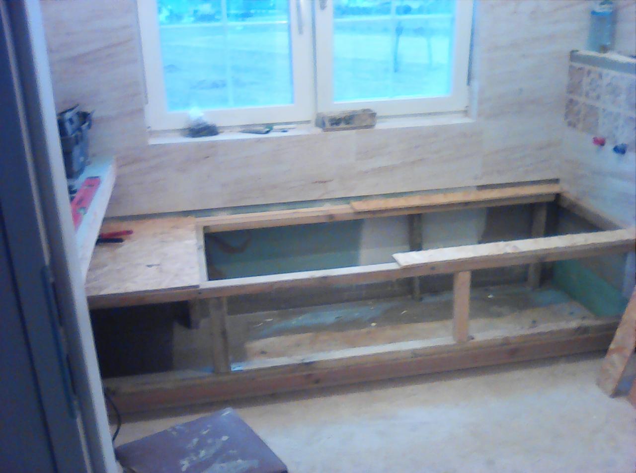 Wnętrze łazienki w trakcie remontu: drewniana konstrukcja wanny w obudowie z płytek, widok na okno i narzędzia na półce.