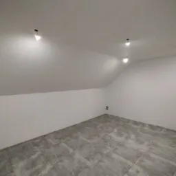 Poddasze z wykończonymi ścianami i szarą, kwadratową podłogą z płytek, widoczne punkty oświetleniowe w suficie i gniazdka elektryczne na ścianie.