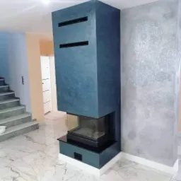 Nowoczesny kominek obudowany w ciemnym odcieniu niebieskiego, z dekoracyjną ścianą w tle z delikatnym wzorem ornamentacyjnym, marmurowa posadzka, schody w tle.