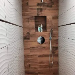 Wykończona kabina prysznicowa z drewnopodobnymi płytkami na ścianie, wnęką na kosmetyki i deszczownicą, kontrastująca z białą, falistą fakturą płytek na sąsiedniej ścianie.