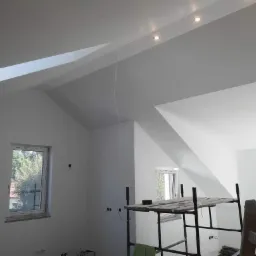 Wnętrze poddasza po remoncie, świeżo pomalowane na biało, widoczne rusztowanie, okno z widokiem na zieleń, oraz oświetlenie punktowe w suficie.