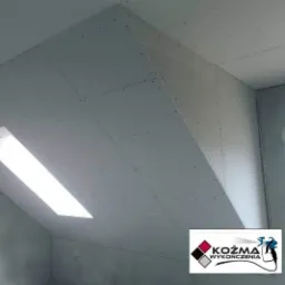 Skośny sufit poddasza z zamontowanymi płytami gipsowo-kartonowymi, widoczne wkręty, okno dachowe i logo firmy w rogu zdjęcia.