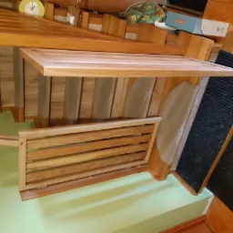 Fragment drewnianej konstrukcji z elementami ażurowymi, prawdopodobnie w trakcie montażu, widoczna na tle zielonej ściany i fragmentu podłogi z ciemną wykładziną.