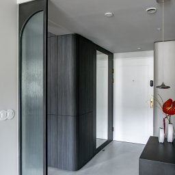 TOMASZ KRALKA FIRMA HANDLOWO USŁUGOWA STOLMAX - Nowoczesna szafa wnękowa z lustrem i zaokrąglonymi narożnikami, wykończona ciemnym drewnem, w minimalistycznym przedpokoju z szarą podłogą i białymi ścianami.