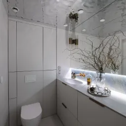 Nowoczesna łazienka z białymi szafkami na wymiar, umywalką i toaletą. Lustro z podświetleniem i dekoracyjnym wazonem. Sufit z efektem lustrzanym.