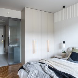 TOMASZ KRALKA FIRMA HANDLOWO USŁUGOWA STOLMAX - Jasna szafa wnękowa z długimi, złotymi uchwytami w sypialni z parkietem w jodełkę. Obok szafy przesuwne drzwi z ryflowanego szkła.