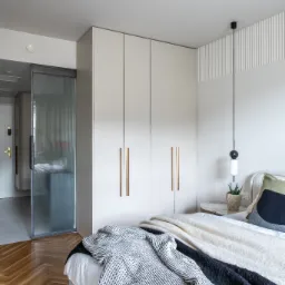 Jasna szafa wnękowa z długimi, złotymi uchwytami w sypialni z parkietem w jodełkę. Obok szafy przesuwne drzwi z ryflowanego szkła.