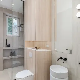 Jasna łazienka z kabiną prysznicową typu walk-in, umywalką nablatową i szafką z drewna. Minimalistyczny design, czarne akcenty, subtelne oświetlenie.