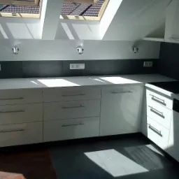 Jasna kuchnia z białymi, błyszczącymi frontami szafek pod skosem dachu, oświetlona naturalnym światłem z okien dachowych.