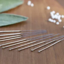 Metalowe igły do akupunktury z spiralnymi uchwytami, obok liść szałwii i białe granulki na drewnianym stole.