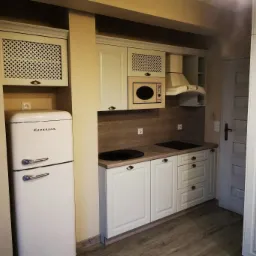 Jasna kuchnia z kremowymi szafkami, retro lodówką i drewnianym blatem. Górne szafki zdobione kratką. Ujęcie prezentuje gotową aranżację.