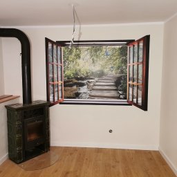 Bartłomiej Kornecki Remonty Kłodzko - Pokój z piecem kaflowym i fototapetą imitującą okno z widokiem na las i wodospad, parkiet na podłodze, jasne ściany.
